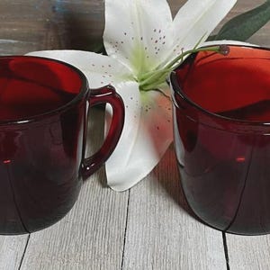 May include: Two matching deep red glass cups, each featuring a rounded body and a single side handle. The glass has a translucent quality, allowing light to pass through the dark crimson material. The cups have smooth, curved rims and bases.
