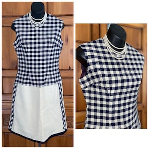 May include: A sleeveless dress featuring a navy blue and white gingham bodice and a solid white skirt panel. The dress is styled with a multi-strand white pearl necklace. The bodice has a high neckline and a fitted silhouette, while the skirt is plain.