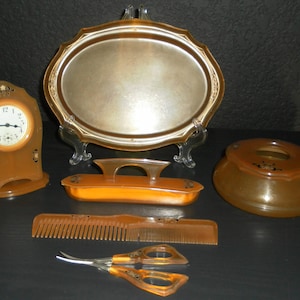 May include: A collection of amber-colored vanity items including an analog clock, an oval tray, a handled brush, a bowl, a comb, and a pair of scissors. Each piece features a translucent, warm brown tone with decorative black and gold accents.