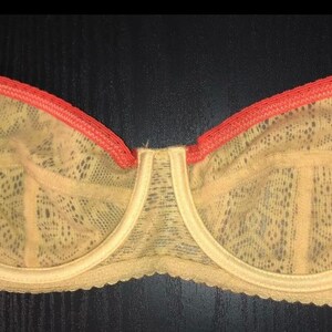 May include: A beige lace bra with a coral trim. The bra features a delicate floral pattern and adjustable straps. The underwire bra has a classic design and is made of a soft, sheer material. The bra is displayed on a dark surface.