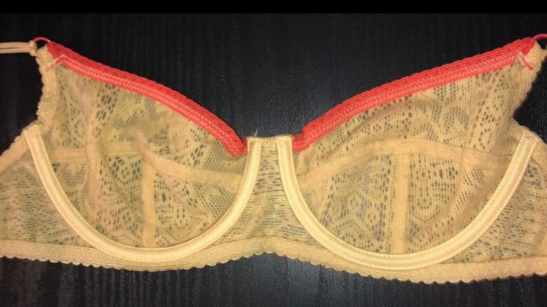 May include: A beige lace bra with a coral trim. The bra features a delicate floral pattern and adjustable straps. The underwire bra has a classic design and is made of a soft, sheer material. The bra is displayed on a dark surface.