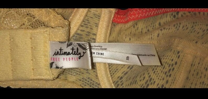 May include: Close-up of a beige bra with lace detailing. The bra features a tag with the words "intimately FREE PEOPLE" in black script. The tag also includes text in other languages. The bra is made in China.