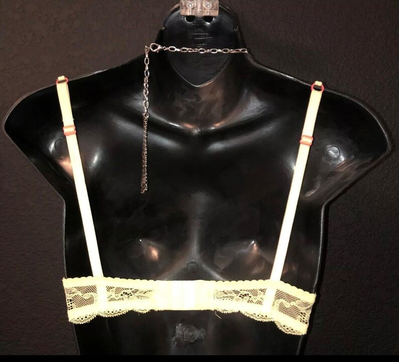 May include: A pale yellow bra with delicate lace detailing along the bottom edge. The bra features thin, adjustable straps and a silver chain necklace. The straps are attached with small, reddish-orange accents. The bra is displayed on a black mannequin.