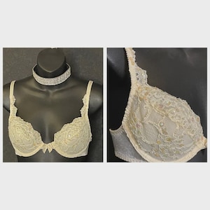 May include: Ivory-colored bra with delicate lace detailing and a small bow. The bra is displayed on a black mannequin, showcasing its intricate design. The lace is embellished with small, iridescent beads.