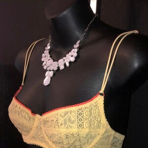 May include: A yellow lace bra with orange trim and thin straps is displayed on a black mannequin. A silver chain necklace with light pink floral and teardrop-shaped pendants is draped around the mannequin's neck.