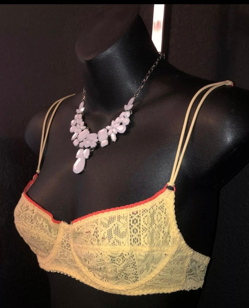 May include: A yellow lace bra with orange trim and thin straps is displayed on a black mannequin. A silver chain necklace with light pink floral and teardrop-shaped pendants is draped around the mannequin's neck.