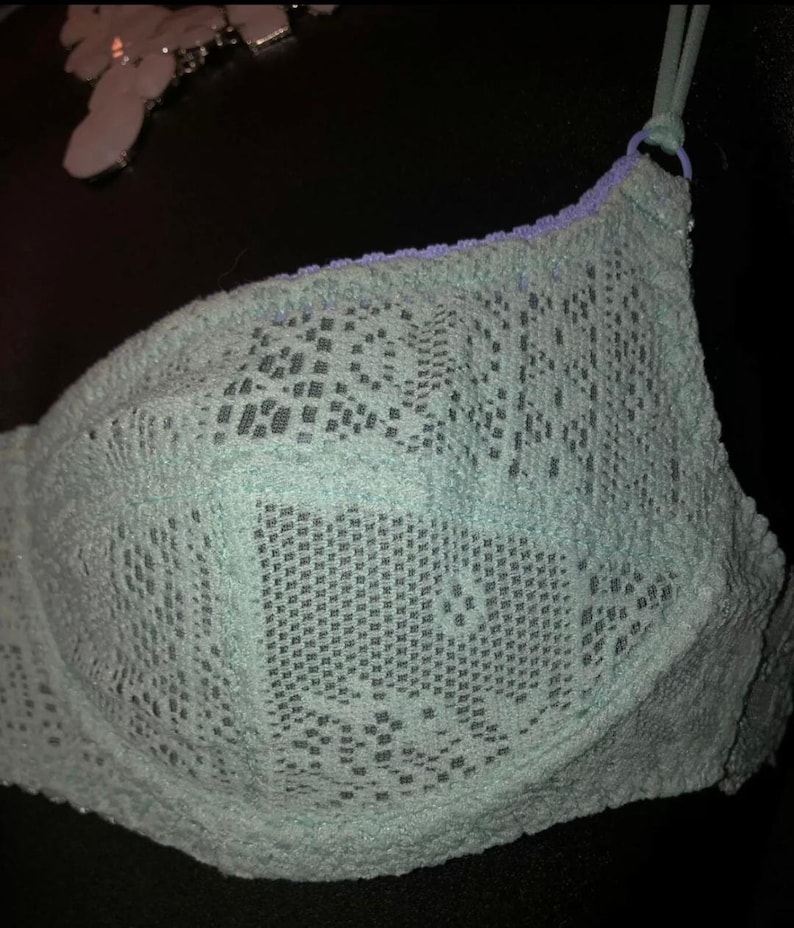 May include: A light blue bra with a delicate lace pattern. The bra features adjustable straps and a contrasting lavender trim along the top edge. The intricate design and soft color create a feminine aesthetic.