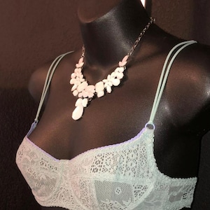 May include: A light green lace bra with thin straps and a matching necklace with white and pink floral accents. The bra is displayed on a black mannequin, showcasing the delicate details of the lingerie.