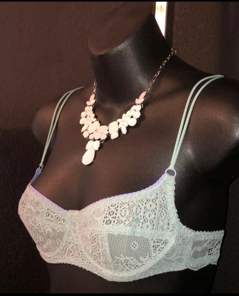 May include: A light green lace bra with thin straps and a matching necklace with white and pink floral accents. The bra is displayed on a black mannequin, showcasing the delicate details of the lingerie.