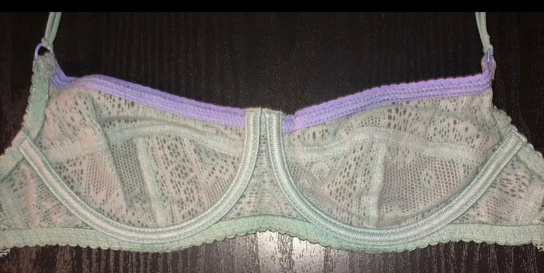 May include: A delicate, lacy bra in shades of pale green and lavender. The underwire bra features a scalloped edge and adjustable straps. The cups are made of a sheer, patterned fabric.