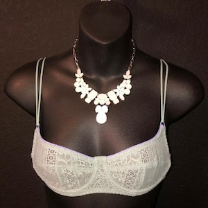May include: A light green lace bra with thin straps is displayed on a black mannequin. A white and pink floral necklace with a pendant is draped over the bra. The bra has a purple trim.