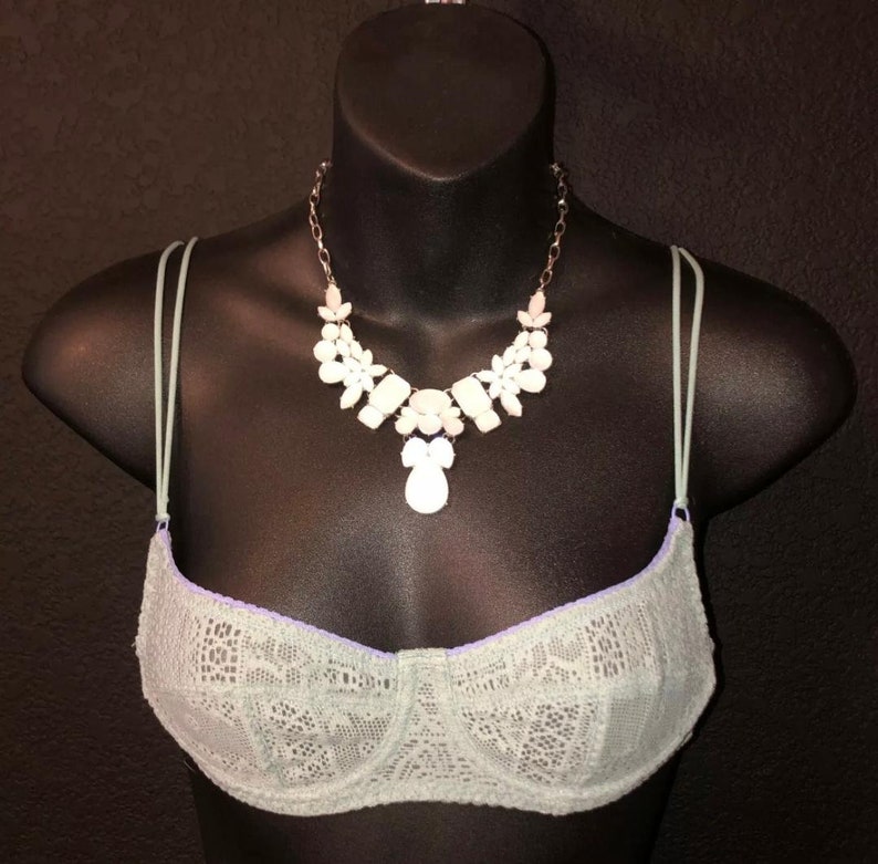 May include: A light green lace bra with thin straps is displayed on a black mannequin. A white and pink floral necklace with a pendant is draped over the bra. The bra has a purple trim.