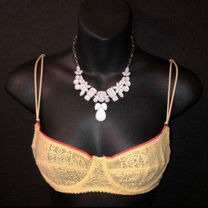 May include: A yellow lace bra with red trim and a white gemstone necklace on a black mannequin. The necklace features a silver chain and white floral and geometric shapes. The bra has thin, beige straps.