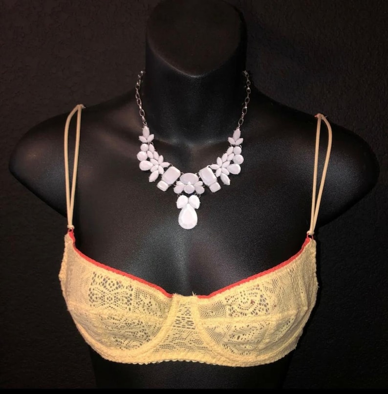 May include: A yellow lace bra with red trim and a white gemstone necklace on a black mannequin. The necklace features a silver chain and white floral and geometric shapes. The bra has thin, beige straps.