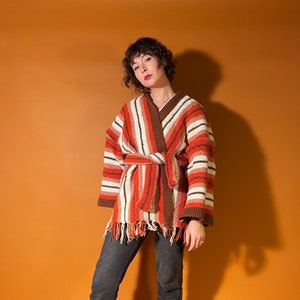 A woman with short brown hair stands in front of an orange backdrop. She is wearing a crocheted orange and brown belted cardigan with black pants and boots