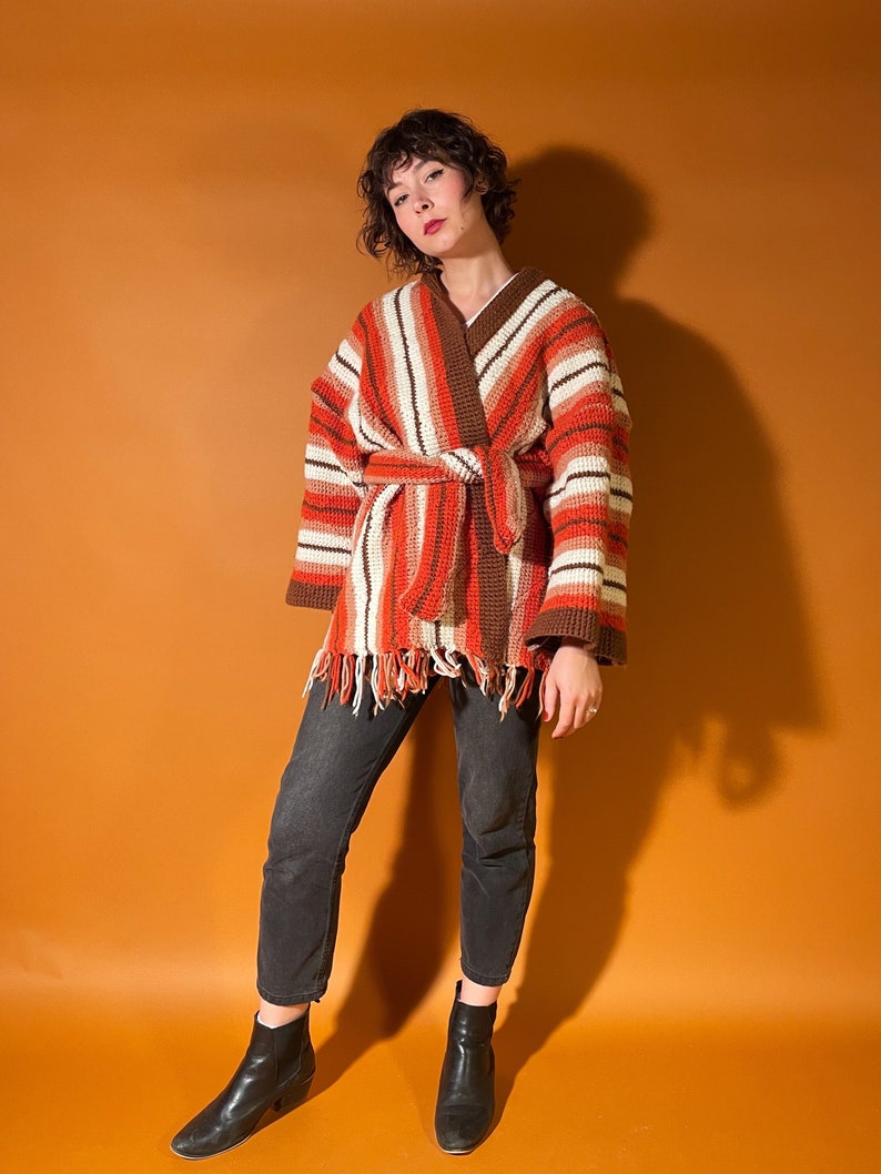 A woman with short brown hair stands in front of an orange backdrop. She is wearing a crocheted orange and brown belted cardigan with black pants and boots