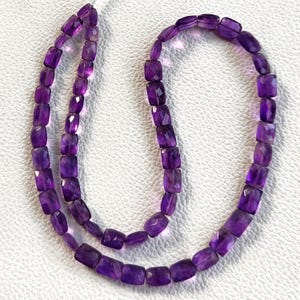Può includere: Una collana di perline di ametista rettangolari e sfaccettate. Le perline sono di un colore viola intenso e infilate insieme. La collana è esposta su una superficie bianca. Le perline hanno una superficie lucida e riflettente.