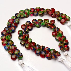 Pode incluir: Um fio de contas de pedras preciosas coloridas com acabamento de manchas douradas. As contas são redondas e têm o verso plano. O fio é terminado com borlas brancas.