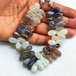 Può includere: Una collana di pietre preziose a forma di goccia in una sfumatura di colori, tra cui bianco, grigio, marrone e nero. Le pietre preziose sono infilate con un cordoncino bianco. Le pietre sono esposte su una mano.