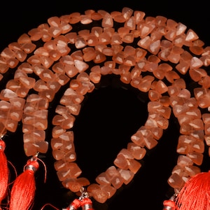 May include: A strand of peach-colored gemstone beads with a faceted, square shape. The beads are strung on a silver wire and have red tassels at each end.