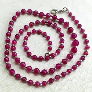 Puede incluir: Un collar con cuentas redondas de color magenta vibrante, ensartadas con pequeñas cuentas plateadas. El collar tiene un cierre plateado y se muestra sobre una superficie clara. Las cuentas varían en tamaño, creando un efecto de capas.