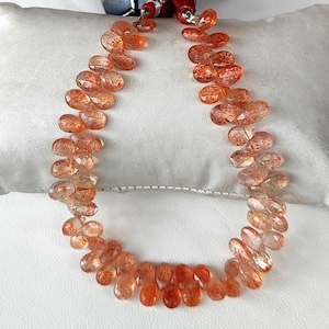 Op de afbeelding: Een ketting bestaande uit gefacetteerde, druppelvormige zonnesteen kralen. De kralen hebben een doorschijnende oranje kleur met sprankelende insluitsels. De ketting is geregen op een rood koord en ligt op een wit oppervlak.