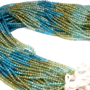 Op de afbeelding: Een streng facetgeslepen edelsteen kralen in tinten blauw en groen. De kralen zijn klein en rond en geregen aan een wit koord. De kralen zijn gerangschikt in een verloop, met de blauwe kralen aan het ene uiteinde en de groene kralen aan het andere uiteinde.