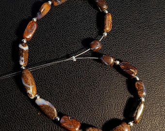 May include: A necklace featuring brown and tan oval-shaped beads with silver-colored spacers. The beads have a marbled appearance with white and blue accents. The necklace is strung on a thin, clear cord.