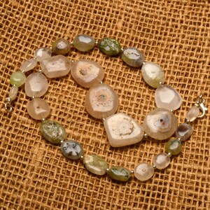 May include: A necklace featuring a variety of polished, irregularly shaped stones in shades of beige, green, and white. The stones are strung together with silver-colored metal links and a clasp. The necklace is displayed on a textured, brown surface.