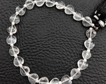May include: A clear quartz beaded bracelet featuring heart-shaped beads strung together with white beads. The bracelet has a black tassel at the end. The beads are transparent and reflect light, creating a sparkling effect.