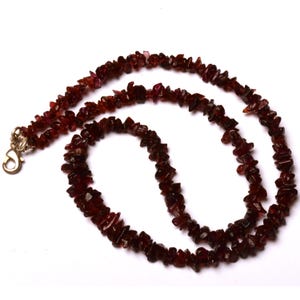 Puede incluir: Un collar hecho de pequeñas piedras de granate rojo oscuro de forma irregular. Las piedras están ensartadas, formando una hebra continua. El collar tiene un cierre dorado.