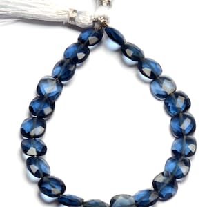 Puede incluir: Un collar de cuentas azules ovaladas y facetadas. Las cuentas están ensartadas, formando un bucle. Borlas blancas con detalles metálicos plateados están unidas a la parte superior. Las cuentas son translúcidas.