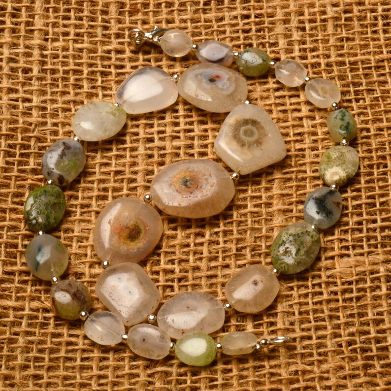 May include: A necklace composed of irregularly shaped, polished gemstones in shades of beige, green, and gray. The stones are strung together with small silver-colored beads and a clasp. The necklace is displayed on a textured, brown surface.