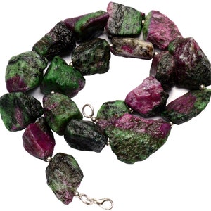 Peut inclure: Un collier composé de perles de pierres précieuses vertes et roses. Les perles sont de forme irrégulière et ont une texture rugueuse et naturelle. Le collier est enfilé sur une chaîne en argent avec un fermoir en argent.