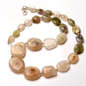 May include: A necklace featuring a variety of irregularly shaped, polished stone beads in shades of beige, green, and white. The beads are strung together with small gold-colored beads and a gold-colored clasp.