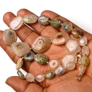 May include: A necklace composed of irregularly shaped, polished agate beads in shades of beige, cream, and green. The beads are strung together with small silver-colored beads and a clasp. The necklace is displayed on a hand.