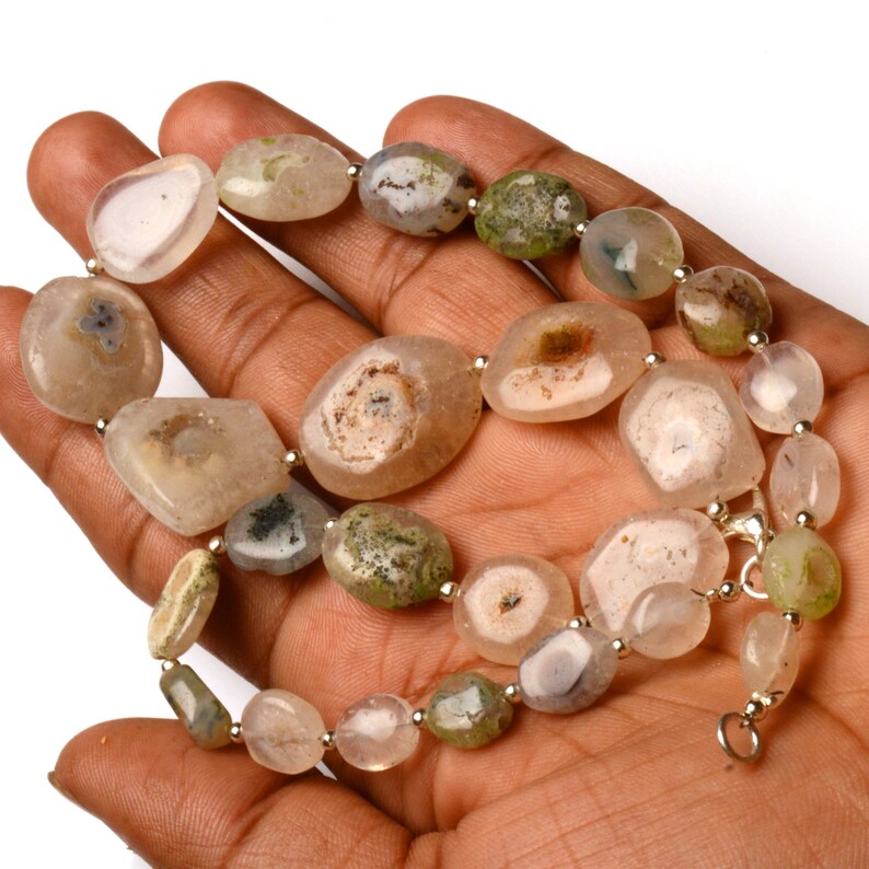 May include: A necklace composed of irregularly shaped, polished agate beads in shades of beige, cream, and green. The beads are strung together with small silver-colored beads and a clasp. The necklace is displayed on a hand.
