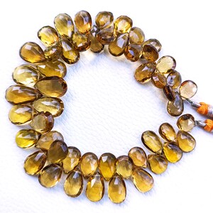 May include: A strand of golden brown teardrop-shaped gemstone beads. The beads are faceted and have a translucent appearance.