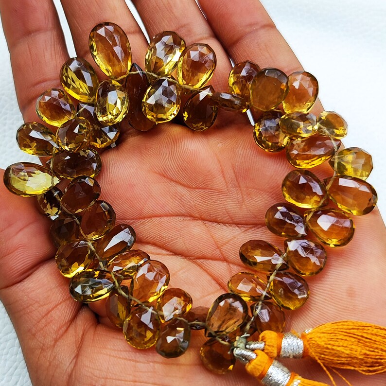 May include: A strand of faceted, golden brown gemstone beads, strung on a thin wire. The beads are oval-shaped and have a faceted, teardrop-like appearance. The strand is finished with two tassels.