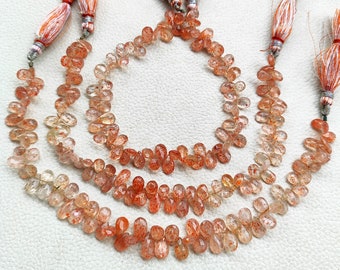 May include: Three strands of sunstone beads, each strand is composed of small, teardrop-shaped beads with a translucent orange color and flecks of red and gold. The beads are strung on a cord with tassels at each end.