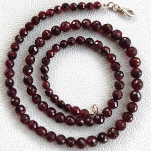 Peut inclure: Un collier composé de pierres précieuses rouge foncé à facettes. Le collier est composé de nombreuses perles rondes et polies enfilées ensemble. Les perles sont d'un rouge bordeaux profond et ont une surface brillante et réfléchissante. Il est sécurisé par un fermoir argenté.