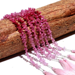 Puede incluir: Una hebra de cuentas de piedras preciosas rosas y rojas, con un efecto de color degradado, sobre una superficie de madera. Las cuentas son pequeñas y tienen una forma ligeramente irregular. La hebra está acabada con un borla.