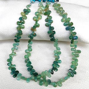 May include: Three strands of green gemstone beads. The beads are teardrop-shaped and vary in shades of green. The strands are strung together with a turquoise-coloured string.