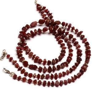 May include: A multi-strand necklace featuring raw, deep red garnet gemstones. The necklace is strung with small, silver-coloured beads and has a silver-toned clasp. The gemstones have a rough, natural texture and vary in size.