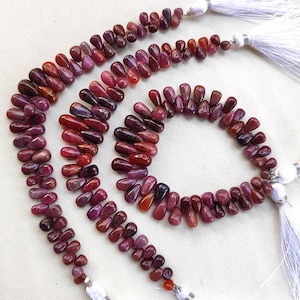 May include: Three strands of red and pink gemstone beads in a teardrop shape. The beads are strung on a white cord with tassels.