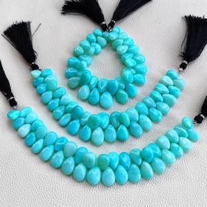 May include: Three strands of bright turquoise, teardrop-shaped beads arranged in a circular pattern and two curved lines. Each end of the bead strands is finished with a black thread tassel secured by a small silver-toned metal cap.