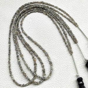 May include: A long strand of raw, uncut grey diamonds arranged in multiple loops. The necklace features small, irregular, textured stone beads in shades of charcoal and silver, finished with two black tassels attached by silver-toned metal connectors.