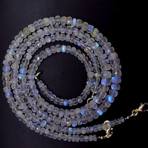 Op de afbeelding: Een lange streng witte maansteen kralen met iriserende blauwe en groene flitsen. De kralen zijn gefacetteerd en hebben een licht rechthoekige vorm. De streng is geregen op een zilveren ketting met een karabijn sluiting.