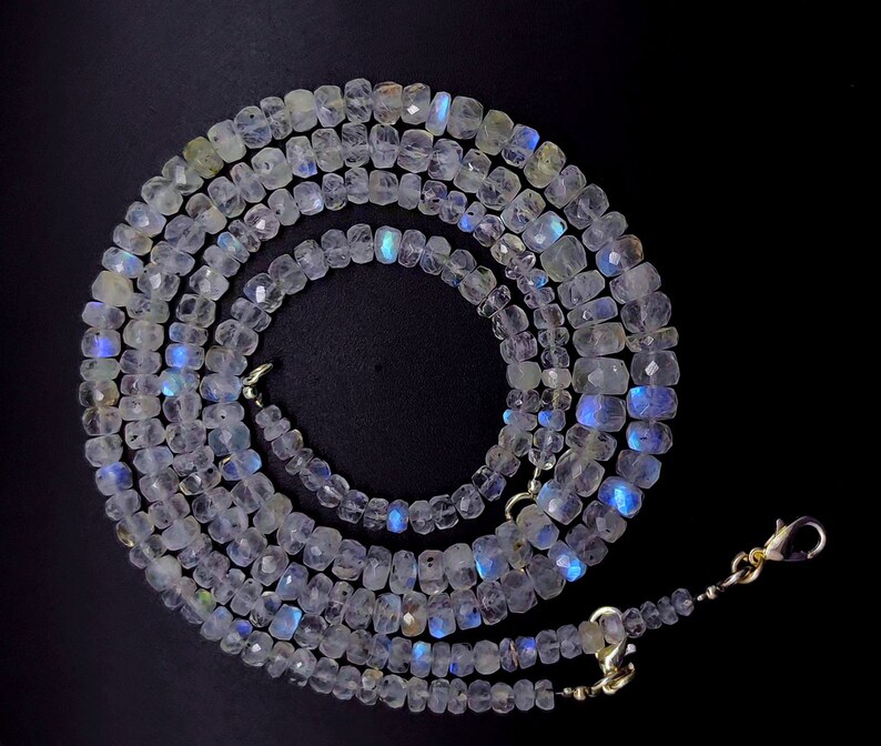 Op de afbeelding: Een lange streng witte maansteen kralen met iriserende blauwe en groene flitsen. De kralen zijn gefacetteerd en hebben een licht rechthoekige vorm. De streng is geregen op een zilveren ketting met een karabijn sluiting.