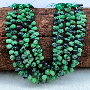 Peut inclure: Gros plan sur plusieurs brins de perles de pierres précieuses vertes et noires. Les perles sont en forme de goutte d'eau et enfilées ensemble, avec quelques perles rouges parsemées. Le fond est une surface en bois brun foncé.